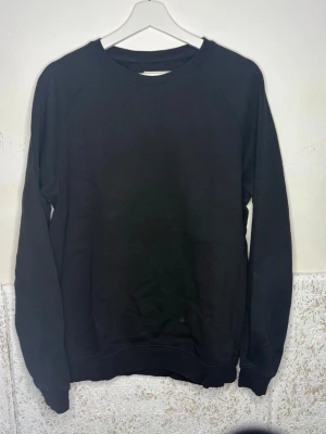 Svart basic sweatshirt - En svart basic sweatshirt med rund hals och långa ärmar. Tröjan har en enkel och clean design utan tryck eller detaljer, vilket gör den lätt att matcha med allt. Tillverkad i mjukt material för en bekväm känsla. Perfekt för dig som gillar minimalistisk stil.