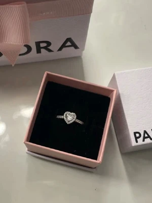 Pandora silverring med hjärta - Säljer en elegant silverring från Pandora med ett gnistrande hjärta i mitten, dekorerad med små klara stenar runt om. Ringen har en tunn, glittrande ringskena och levereras i original ask. Perfekt för dig som gillar romantiska detaljer.