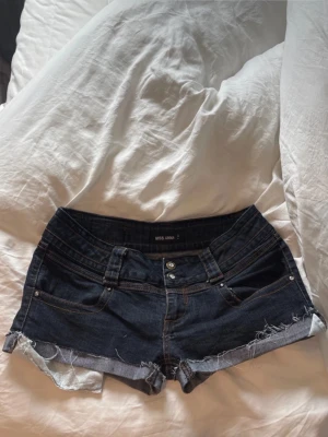 Mörkblå jeansshorts från Miss Anna - 40 cm midja 16 cm rise  Storlek 42 Säljer ett par mörkblå jeansshorts från Miss Anna med råa, fransiga kanter och klassisk femficksdesign. Shortsen har låg midja och silverfärgade knappar. Perfekta för sommardagar och festivaler.