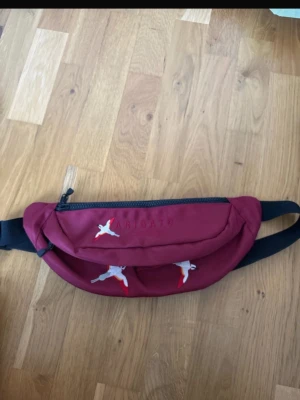 Vinröd magväska från Axel Arigato - Snygg vinröd magväska från Axel Arigato med broderade fåglar i vitt och rött. Väskan har ett stort fack med dragkedja och justerbart svart band. Perfekt accessoar för att bära det viktigaste med stil.