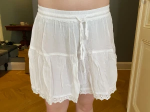 Vit kjol - Söt vit kortkjol med volang och dekorativ spetskant längst ner. Kjolen har resår i midjan och knytband framtill för en bekväm passform. Perfekt för varma dagar och enkel att matcha med olika toppar.