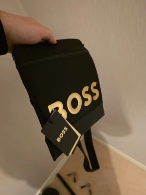 Svart axelväska från BOSS - Snygg svart axelväska från BOSS med stort guldfärgat BOSS-tryck på framsidan. Väskan har justerbar axelrem och dragkedja upptill. Tillverkad i ett slätt syntetmaterial som ger en modern look. Perfekt för att bära det viktigaste och matcha till streetwear.