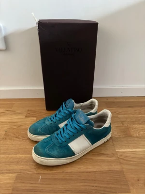 Blåa sneakers från Valentino - Snygga blåa sneakers från Valentino med vita detaljer och vit sula. Skorna har en klassisk låg siluett och är tillverkade i mocka och läder. Perfekta för dig som vill ha en stilren och exklusiv look.