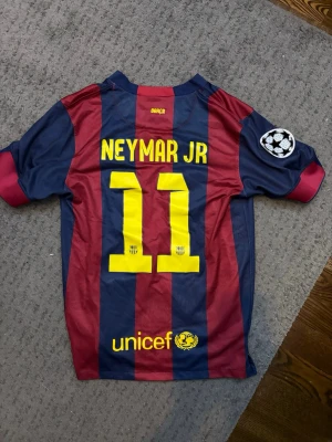Barcelona Neymar Jr matchtröja 2015 - Säljer en FC Barcelona matchtröja från Nike med Neymar Jr och nummer 11 på ryggen. Tröjan har breda blå och vinröda ränder, Champions League-märke på ärmen och tryck från Qatar Airways och UNICEF. Perfekt för dig som älskar fotboll och Barca!