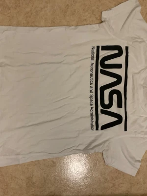 Vit NASA t-shirt med tryck - Säljer en vit t-shirt med stort svart NASA-tryck på framsidan och texten 'National Aeronautics and Space Administration' längs loggan. Klassisk passform och korta ärmar. Perfekt för dig som gillar rymden och vill ha en clean look.