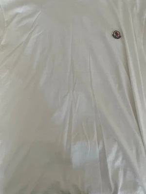 Vit Moncler t-shirt med logga - Säljer en klassisk vit t-shirt från Moncler med den ikoniska loggan broderad på bröstet. T-shirten har rund hals och korta ärmar, perfekt för en clean och stilren look. Tillverkad i mjuk bomull för maximal komfort. Strl L/Xl