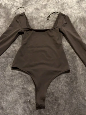 Brun  långärmad body - Säljer en brun långärmad body med djup urringning och tunna axelband. Bodyn har en enkel och stilren design, perfekt att matcha med jeans eller kjol. Materialet känns mjukt och stretchigt för en bekväm passform.