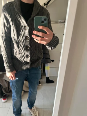 Grå kabelstickad kofta med sjalkrage - Säljer en grå kabelstickad kofta från Tommy Hilfiger med sjalkrage och stora knappar framtill. Koftan har grovstickat mönster, ribbade muddar och en avslappnad passform. Perfekt för lager på lager och riktigt snygg till jeans.