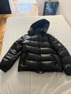 Svart dunjacka från Moncler - Snygg svart dunjacka från Moncler med glansig finish, huva och två dragkedjeförsedda fickor framtill. Jackan har en klassisk Moncler-logga på ärmen och breda kanaler fyllda med dun för extra värme. Perfekt för kalla vinterdagar.