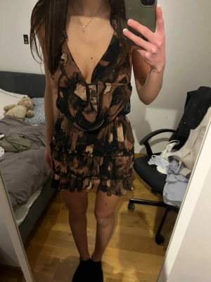 Brun och svart omlottklänning - Säljer en kort brun och svart omlottklänning med volangdetaljer och djup v-ringning. Klänningen har öppen rygg med tunna axelband och ett unikt abstrakt mönster. 