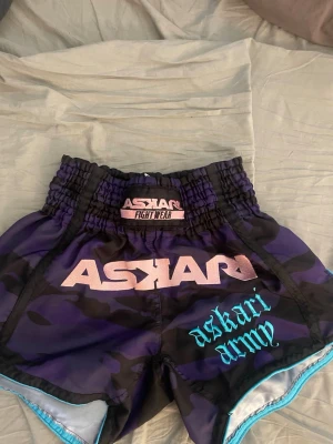 Lila kamouflage fight shorts Askari - Säljer ett par lila och svarta kamouflage fight shorts från Askari. De har bred resår i midjan, ljusblå detaljer och tryck med 'ASKARI' och 'askari army' på framsidan. Perfekta för kampsport eller träning.