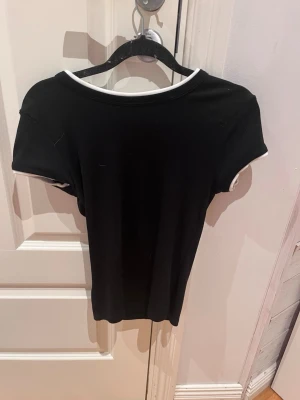 Svart t-shirt med vit kant - En stilren svart t-shirt med korta ärmar och vit kant runt halsringningen och ärmsluten. Modellen är figurnära och enkel, perfekt för en clean och modern look. Materialet ser ut att vara mjukt och stretchigt, troligen bomull eller bomullsblandning.
