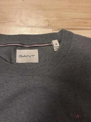 Grå rundhalsad tröja från GANT - Klassisk grå tröja från GANT med rund halsringning och diskret broderad logga på bröstet. Tillverkad i mjukt material som känns skönt mot huden. Perfekt basplagg för en clean och stilren look. Pris kan diskuteras