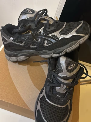 Svarta och silver Asics sneakers - Snygga Asics sneakers i svart och silver med meshpaneler för bra ventilation och detaljer i syntetmaterial. Skorna har klassisk snörning och en chunky sula som ger grym streetstil. Perfekta för dig som gillar sportig look och vill ha bekväma sneakers till vardags. Använda 1 gång 