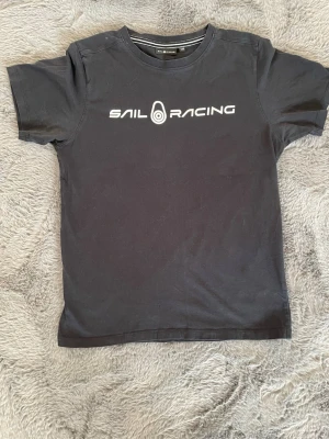 Svart Sail Racing t-shirt - !DET STÅR 170cm MEN ÄR MINDRE I STORLEK! Svart t-shirt från Sail Racing med vit logotyptryck på bröstet och en mindre logga i nacken. Klassisk rund hals och korta ärmar. Tillverkad i mjuk bomull för en skön känsla och enkel stil.