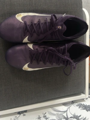 Lila Nike fotbollsskor med dobbar - Säljer ett par lila Nike fotbollsskor med vita detaljer och rosa dobbar. Skorna har klassisk snörning och är tillverkade i syntetmaterial för bra grepp och kontroll på planen. Perfekta för dig som vill sticka ut på matchen! Storlek 43
