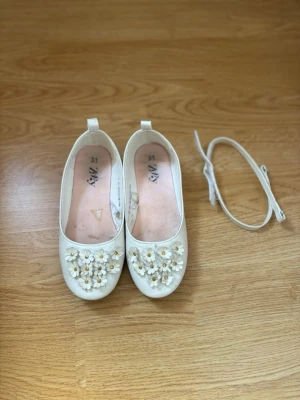 Vita ballerinaskor med blommor  - Säljer ett par vita ballerinaskor från Zaxy med söta 3D-blommor och pärldetaljer framtill. Skorna har rund tå och medföljande remmar som kan fästas runt ankeln. 