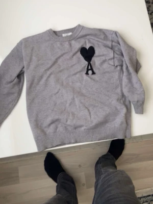 Ami paris tröja - Säljer en grå stickad tröja med svart hjärta och bokstaven A broderat på bröstet. Tröjan har rund halsringning, ribbade muddar och nederkant. Perfekt för dig som gillar stilrena plagg med en twist.