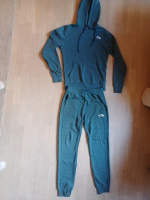 Grönt loungewear-set från The North Face - Mysigt loungewear-set i grönt från The North Face. Setet består av en hoodie med dragsko och magficka samt matchande byxor med resår i midjan och muddar vid bensluten. Båda delarna har The North Face-logga tryckt på bröstet och benet. Perfekt för chill dagar hemma.