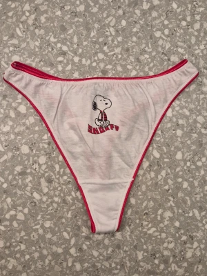 Vita Snoopy-trosor med rosa kant - Snygga vita stringtrosor med hög midja och rosa kantband. Framtill finns ett tryck av Snoopy i röd halsduk och texten 'Snoopy'. Tillverkade i 100% bomull för en mjuk och skön känsla. Perfekta för dig som gillar gulliga och lekfulla detaljer i din underklädesgarderob. Finns i storlek 34/36, 38/40 eller 42/44. Äkta Y2K från restlager från 2000-talet. Bundlerabatt vid flerköp.