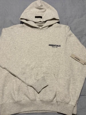 Grå Essentials hoodie från Fear of God - Snygg ljusgrå hoodie från Essentials Fear of God med svart logga på bröstet och ryggen. Klassisk passform med huva och ribbade muddar. Perfekt för en avslappnad och trendig streetwear-look. Materialet är mjukt och bekvämt, troligtvis bomullsblandning. Och ljusgrå mjukisbyxor från Essentials Fear of God med svart logga på benet. De har resårmidja med snörning och är gjorda i mjukt bomullsmaterial. Perfekta för en chill och trendig look.
