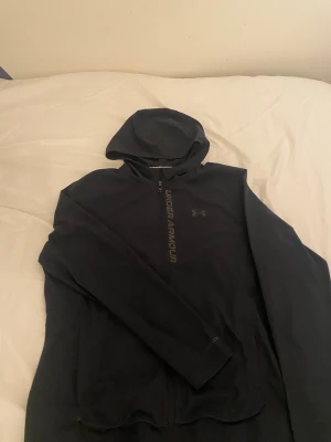 Svart hoodie från Under Armour - Svart hoodie från Under Armour med dragkedja och huva. Lätt och ventilerande material, perfekt för lager-på-lager. Logga på bröstet och tryck längs dragkedjan ger en sportig vibe. Sitter snyggt och är enkel att matcha.