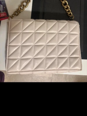 Beige quiltad handväska från Zara - Snygg beige handväska från Zara med quiltat mönster och grov guldfärgad kedja som axelrem. Väskan är rektangulär och tillverkad i ett glansigt konstlädermaterial. Perfekt för att lyfta vilken outfit som helst.
