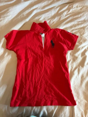 Röd pikétröja från Ralph Lauren - Röd pikétröja från Ralph Lauren med klassisk krage, korta ärmar och broderad marinblå logga på bröstet. På ena ärmen finns en stor broderad siffra 3 och texten 'Polo'. Perfekt för en sportig och stilren look. Vet ej om den är äkta 