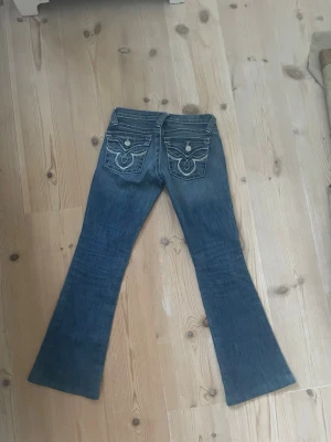 Blå bootcut jeans med broderade fickor - Dom sitter väldigt lågt på. Snygga blå bootcut jeans med broderade bakfickor och dekorativa knappar. Jeansen har klassisk femficksdesign och markerade sömmar..