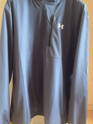 Blå vindjacka från Under Armour - Blå vindjacka från Under Armour med hel dragkedja och en vertikal bröstficka med svart dragkedja. Jackan har lång ärm, hög krage och är gjord i ett lätt, tekniskt material som skyddar mot vind. Perfekt för träning utomhus eller när du vill ha en sportig look.