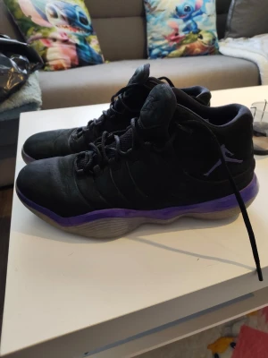 Svarta Air Jordan sneakers med lila sula - Snygga svarta Air Jordan sneakers med lila detaljer på sulan och klassisk Jumpman-logga på sidan. Skorna har högt skaft, svarta skosnören och en robust, vågformad sula. Materialet är en mix av mocka och textil för en cool och sportig look.