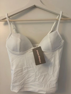Vit top med tunna axelband Missguided - Snygg vit top från Missguided med tunna, justerbara axelband och vadderade kupor. Mjukt och stretchigt material som sitter bekvämt mot kroppen. Perfekt att styla under skjortor eller tröjor. Märkesdetalj baktill.