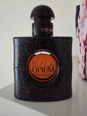 Yves Saint Laurent Black Opium parfym - Black Opium från Yves Saint Laurent är en ikonisk parfym i en glittrig svart flaska med rund orange detalj. Flaskan har en modern, stilren form och känns lyxig med sin svarta och orange färgkombination. Perfekt för dig som gillar eleganta och trendiga dofter. Använd lite, det syns hur mycket parfymen finns kvar. 30 ml från början. 