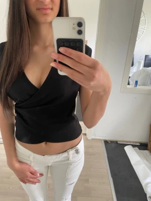 Svart omlottopp med gulddetalj - Snygg svart omlottopp med v-ringning och guldfärgad knappdetalj i sidan. Toppen är croppad och har en figurnära passform utan ärmar. Perfekt för dig som vill ha en stilren och trendig look.