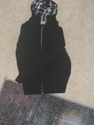 Burberry koft - Cool svart hoodie med hel dragkedja och rutigt foder i huvan. Huvan har snören och fodret har svart, vitt och rött rutmönster. Tröjan har ribbade muddar och är perfekt för en avslappnad look. Materialet ser ut att vara mjukt och bekvämt.