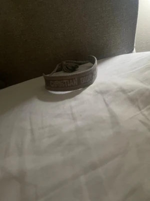 Beige broderat armband från Dior - Snyggt beige armband från Christian Dior med broderad logotyp och justerbar knytning. Armbandet är gjort i textil och har en trendig, avslappnad look som passar perfekt till din outfit.