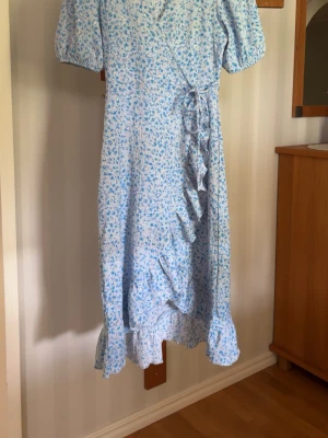 Blommig omlottklänning i blått - Söt långklänning med ljusblått blommönster och volangkant. Klänningen har puffiga korta ärmar, omlottmodell med knyt i midjan och är tillverkad i ett lätt bomullstyg. Perfekt för sommarens alla tillfällen.