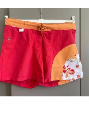 Färgglada badbyxor från Trespass - Säljer ett par badbyxor från Trespass i rött och orange med snörning i midjan. De har en cool detalj med blommigt mönster på ena sidan och en metallöljettdetalj. Perfekta för stranden eller poolen, riktigt sköna och snygga.