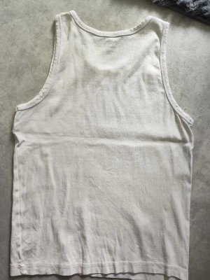 Vit ribbad linnetopp - En klassisk vit ribbad linnetopp i bomull. Modellen är ärmlös med rund halsringning och har en enkel, rak passform. Perfekt att styla till jeans eller shorts för en avslappnad look.