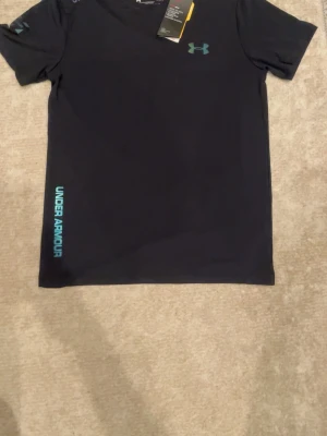 Svart tränings-tshirt från Under Armour - Svart sportig t-shirt från Under Armour med turkos logga på bröstet och text längs sidan. Tillverkad i ett lätt och snabbtorkande material som passar perfekt för träning och aktivitet.