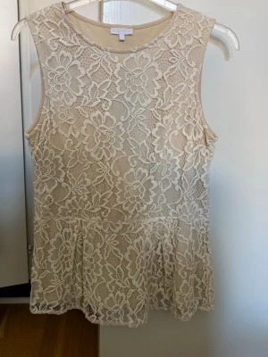 Beige spetstopp från Stockh LM - Superfin beige topp från Stockh LM med blommig spets och ärmlös design. Toppen  har rund halsringning och ett vackert, feminint mönster i spetsmaterial som ger en romantisk känsla. Innerfoder 100% bomull.