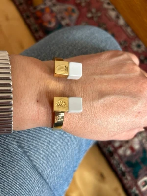 Ioaku armband med vita detaljer - Snyggt armband i guldmetall med två vita fyrkantiga detaljer i ändarna. Ena änden har en liten guldig dekoration. Stilren och modern design som passar perfekt till din accessoar-samling.