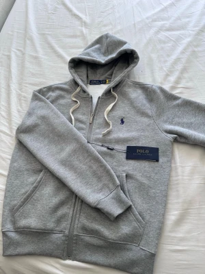 Grå hoodie från Polo Ralph Lauren - Snygg grå hoodie från Polo Ralph Lauren med dragkedja, vit dragsko i huvan och klassisk blå logga broderad på bröstet. Tillverkad i mjuk bomullsblandning och har två fickor framtill. Perfekt för en chill och stilren look.