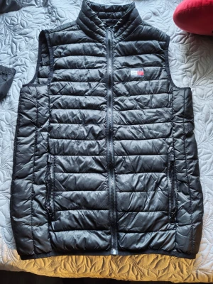 Svart dunväst från Tommy Hilfiger - Snygg svart dunväst från Tommy Hilfiger med klassisk quiltad design och dragkedja framtill. Västen har två praktiska fickor med dragkedja och en ikonisk logga på bröstet. Perfekt för lager-på-lager och streetstyle.
