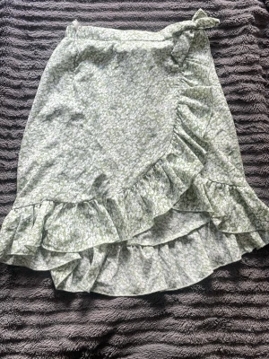 Grön blommig omlottkjol med volang - Söt grön omlottkjol med vitt blommönster och volangkant. Kjolen är perfekt för vår och sommar. Lätt och luftig modell som ger en romantisk vibe.
