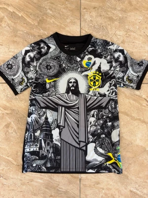 Nike Brasil Jesus tröja - Unik grafisk t-shirt från Nike med Brasil-motiv och stora tryck av Kristusstatyn, änglar och detaljer i svart, vitt och gult. Bröstet har CBF-logga och Nike Swoosh. Tillverkad i lätt och bekväm polyester, perfekt för dig som gillar streetwear och fotbollsinspirerad stil.