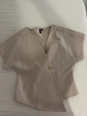 Beige omlottblus från Vero Moda - Stilren beige omlottblus från Vero Moda med korta ärmar och en snygg guldig knapp framtill. Blusen har en djup v-ringning och är tillverkad i ett mjukt, vävt tyg som ger en elegant siluett.