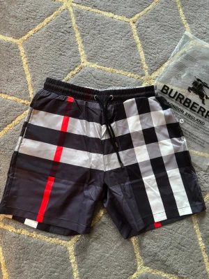 Burberry shorts - Snygga shorts från Burberry med klassiskt rutigt mönster i svart, vitt och rött. Shortsen har elastisk midja med snörning och en bakficka. Perfekta för sommaren och ger en trendig look. Storlek M men rekommenderar för personer med S