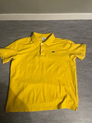 Gul pikétröja från Lacoste - Gul pikétröja från Lacoste med klassisk krage och två knappar framtill. Tröjan har det ikoniska krokodilmärket broderat på bröstet och är tillverkad i bomull. Perfekt för en avslappnad och stilren look.