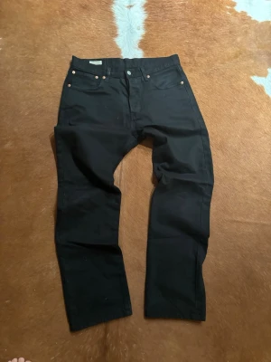Svarta Levi's 501 jeans - Klassiska svarta Levi's 501 jeans med rak passform och fem fickor. Jeansen har knappgylf och den ikoniska läderpatchen bak i midjan. Perfekta för en stilren och tidlös look.